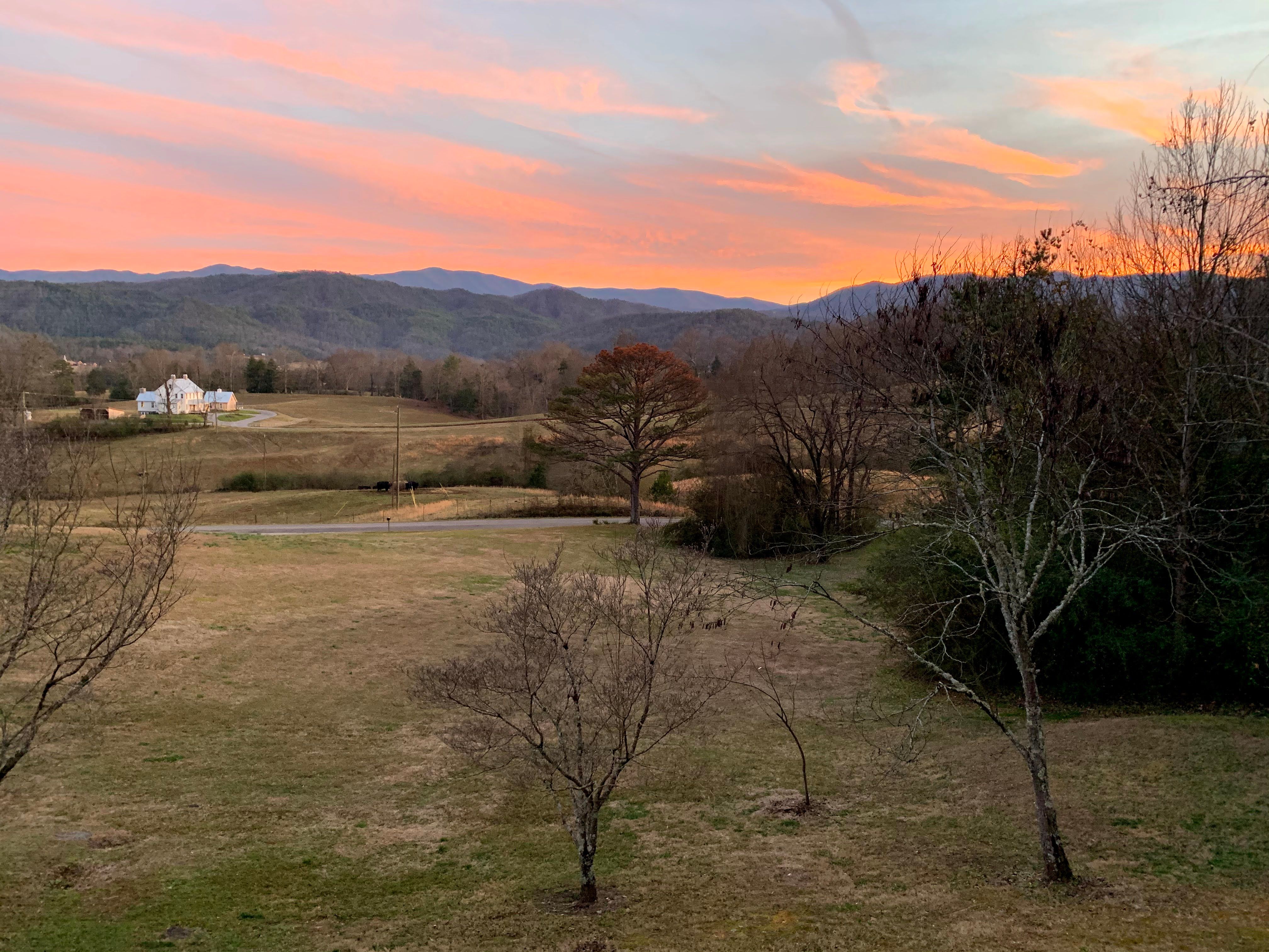 Sunset over the Townsend valley — a pink and orange sky above the layered Smoky Mountain ridgelines, with open pasture and a white farmhouse in the distance